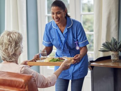 female-care-worker-in-uniform-bringing-meal-on-tray-to-senior-woman-sitting-in-lounge-at-home.jpg